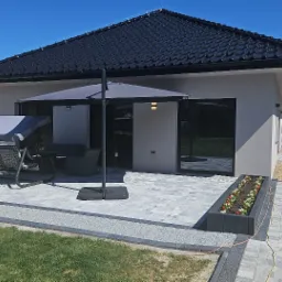 Nowoczesny dom z grafitowym dachem i jasną elewacją. Taras z szarej kostki brukowej, meble ogrodowe i parasol. Widok na zadbany ogród i ogrodzenie.