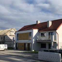 Eco Sense Sp. z o.o. - Nowoczesny dwupiętrowy dom z balkonem i czerwonym dachem, obok drugi dom w trakcie budowy z widoczną konstrukcją dachu, na pierwszym planie ulica i skrzynki elektryczne.