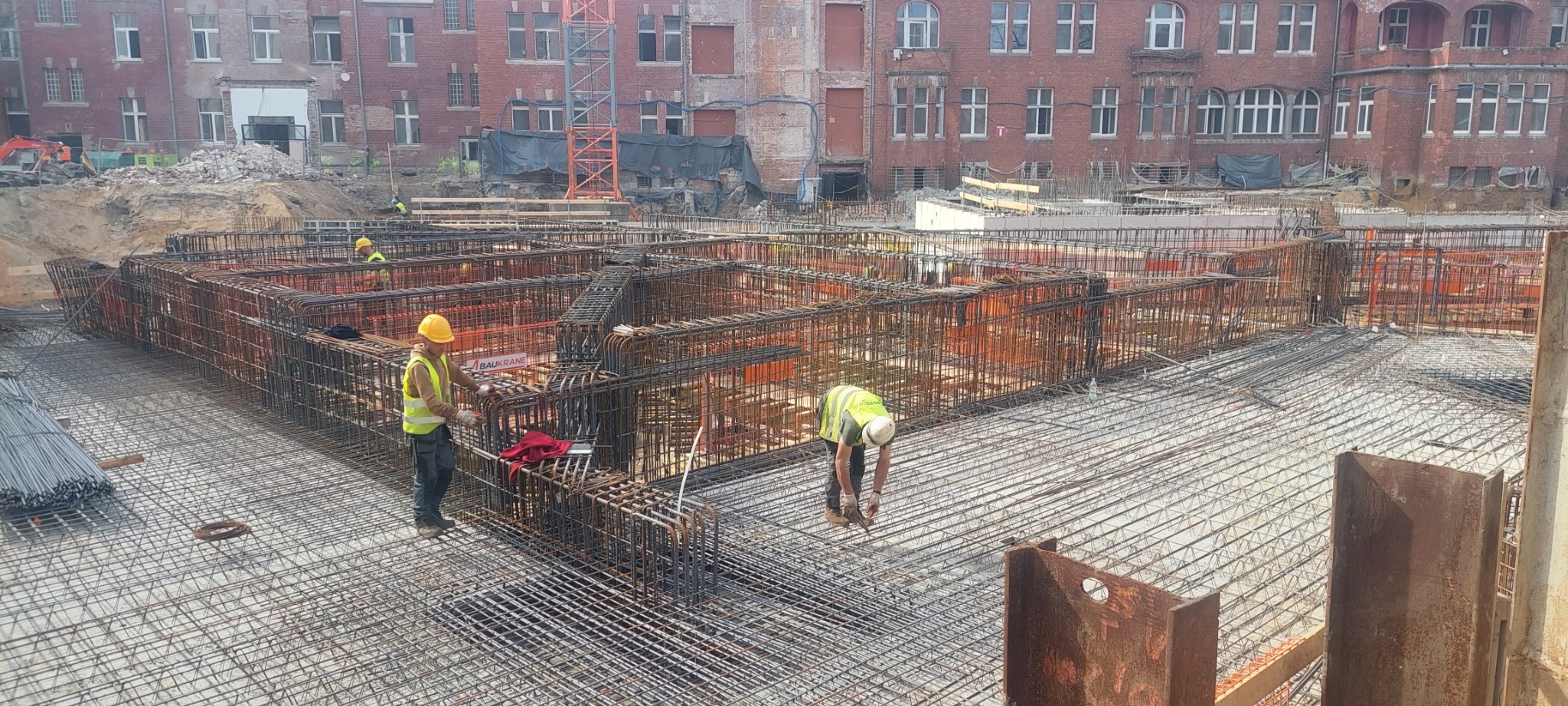 Zbrojenie fundamentów budynku z widocznymi pracownikami w kaskach i kamizelkach odblaskowych, na tle ceglanego budynku i żurawia budowlanego.