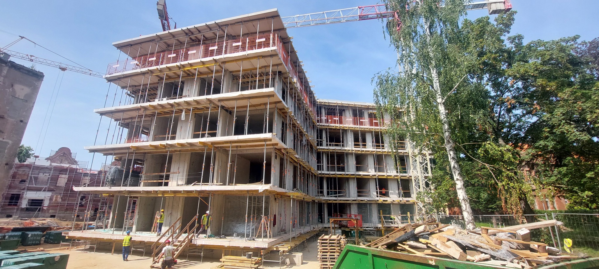 Wznoszenie wielopiętrowego budynku z betonu komórkowego w otoczeniu zieleni i z widocznym żurawiem budowlanym. Na pierwszym planie pracownicy w kaskach.