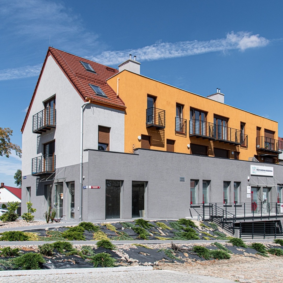 Nowoczesny budynek z elewacją w odcieniach szarości, bieli i żółci, balkony z czarnymi balustradami, czerwony dach i częściowo widoczne niebo.