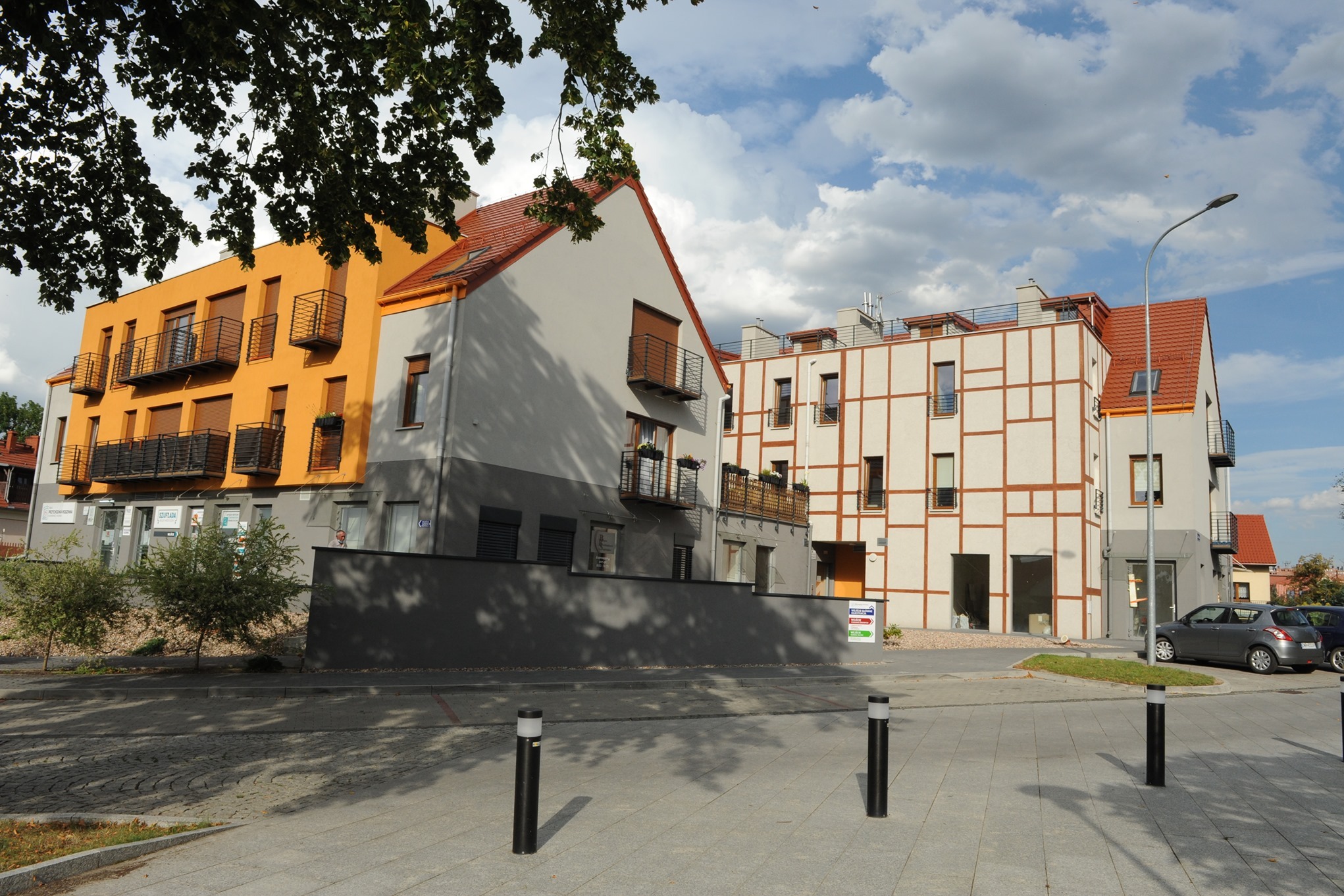 Kompleks budynków o różnorodnej architekturze: pomarańczowa elewacja z balkonami, szara bryła z czerwonym dachem oraz budynek w stylu muru pruskiego z brązowymi belkami.