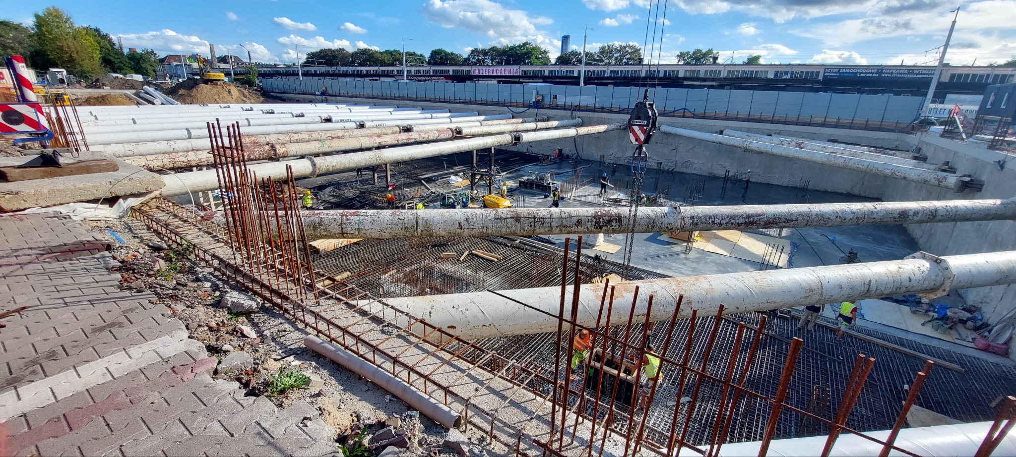 Budowa fundamentów z rurami ochronnymi i zbrojeniami; widoczni pracownicy i dźwig. Etap prac ziemnych i konstrukcyjnych w otoczeniu miejskim.