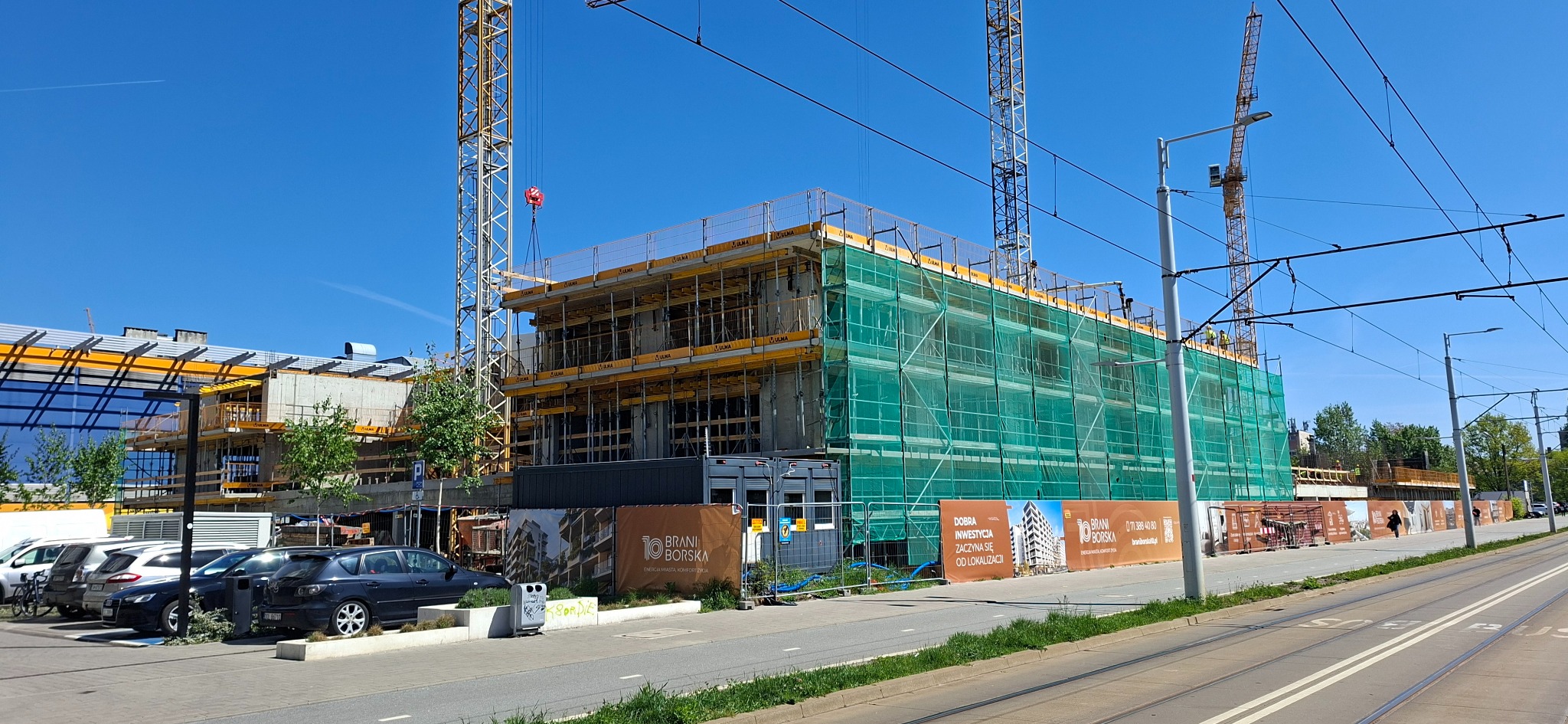Wznoszenie budynku z betonu komórkowego, zabezpieczonego siatką. Na budowie widoczne rusztowania, dźwigi i reklamy inwestycji. Błękitne niebo w tle.
