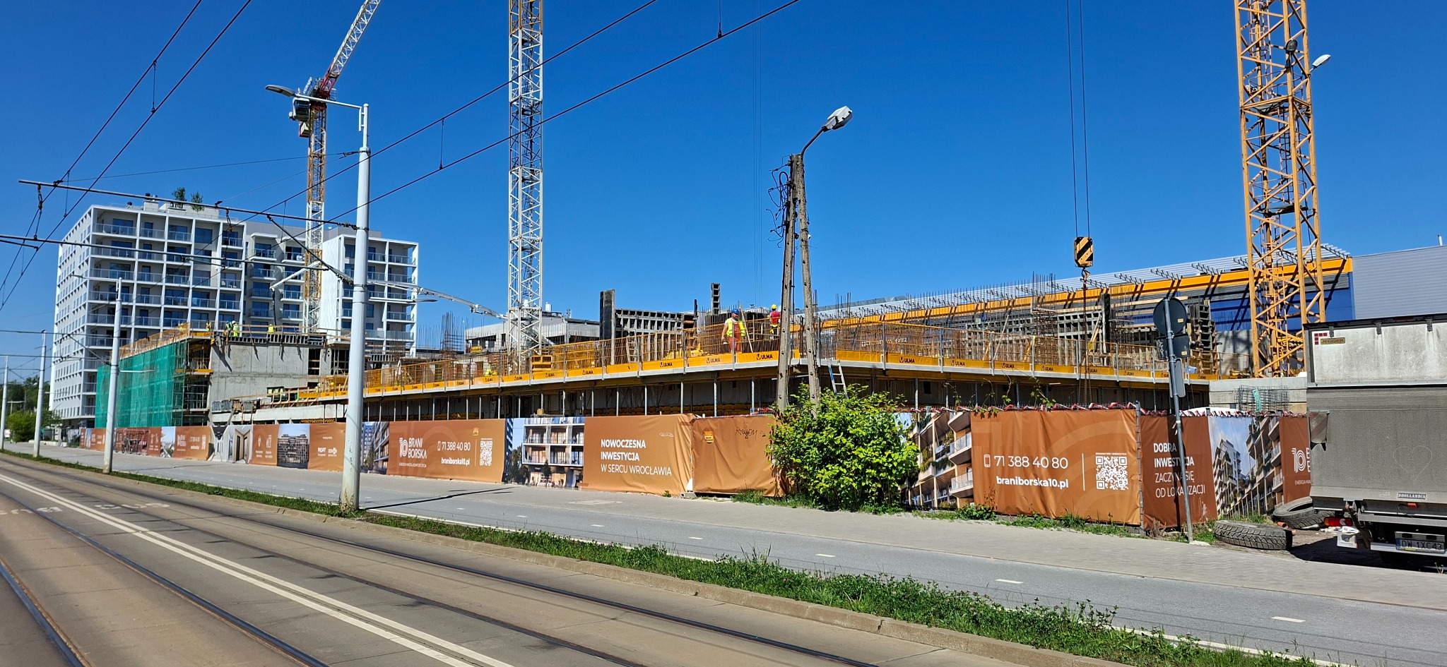 Budowa apartamentowca w słoneczny dzień. Widoczne rusztowania, dźwigi i ogrodzenie z reklamami inwestycji. Ujęcie z poziomu ulicy, z torami tramwajowymi.