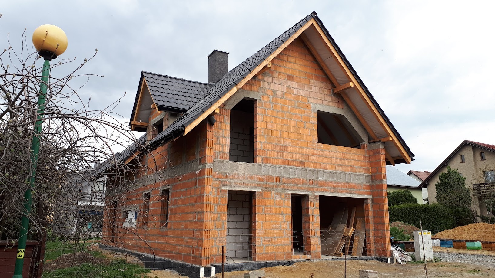 Surowy dom z czerwonej cegły w trakcie budowy, z widocznym kominem i oknami bez szyb. W tle widoczny słup oświetleniowy z kulistą lampą i inne budynki.