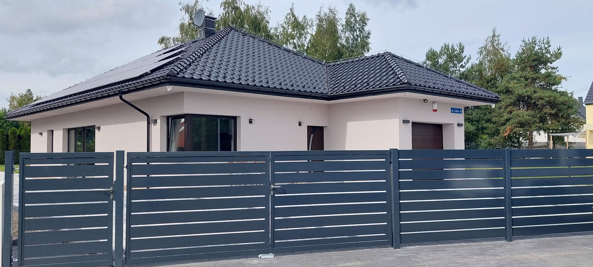 Nowoczesny dom z panelami słonecznymi na dachu, widoczny zza metalowego ogrodzenia z poziomymi szczeblami. Elewacja jasna, dach grafitowy, otoczenie zielone.