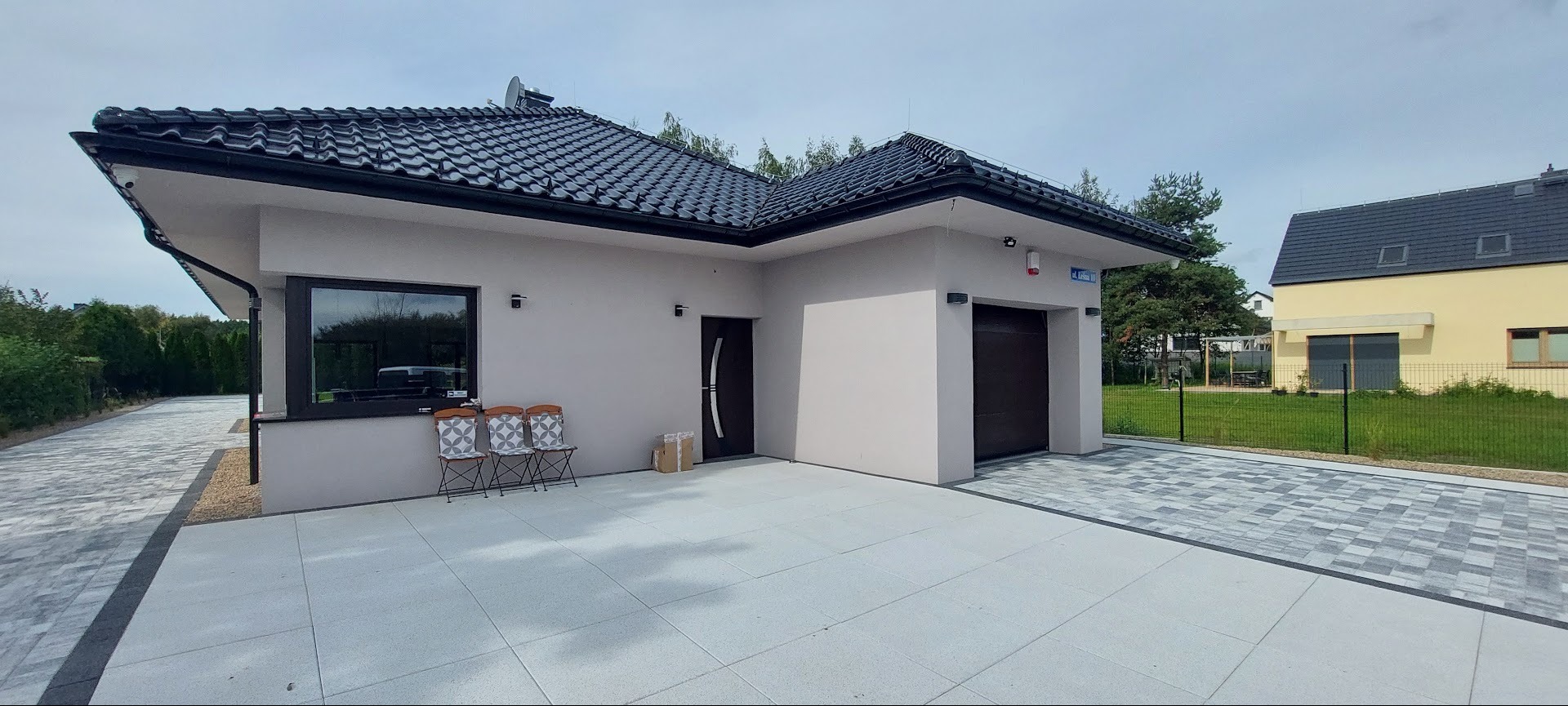 Nowoczesny dom z garażem, szara elewacja, czarny dach z dachówki, kostka brukowa na podjeździe. Widoczne krzesła ogrodowe i paczka. Dom sąsiaduje z innym budynkiem.