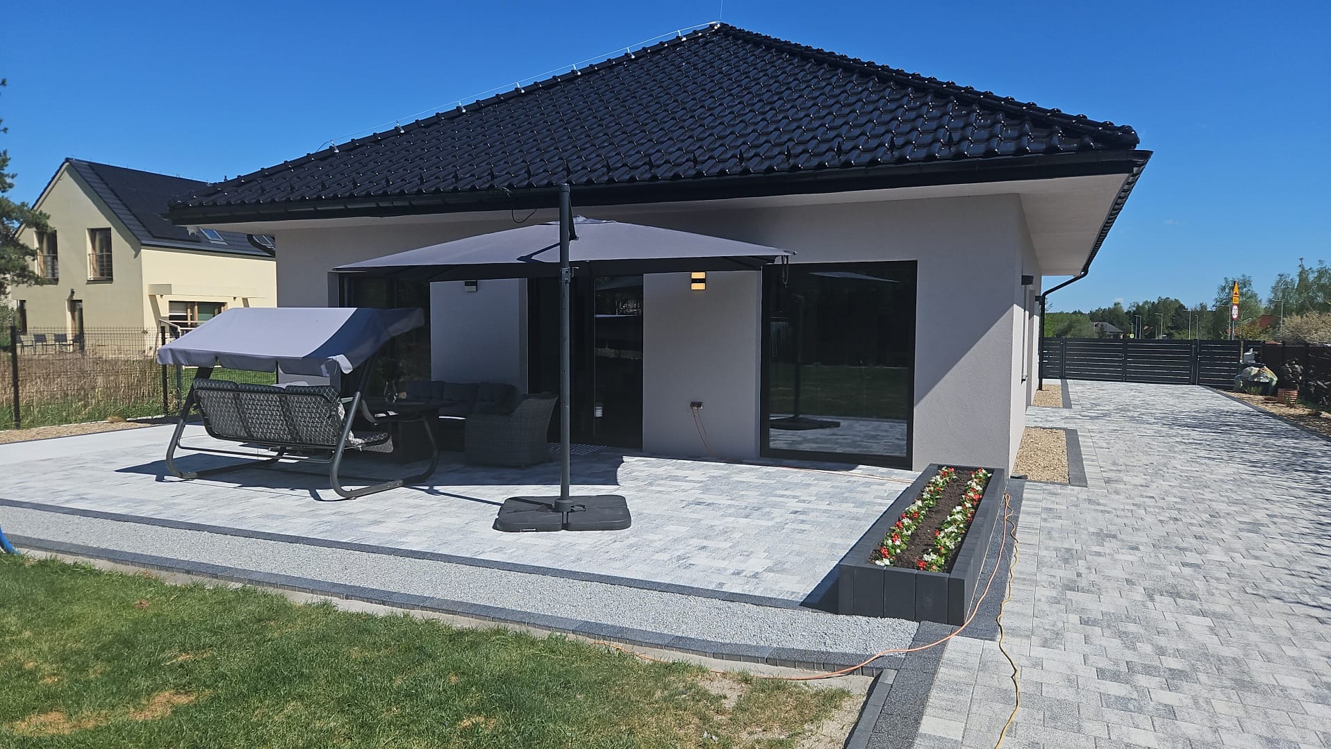 Nowoczesny dom z grafitowym dachem i jasną elewacją. Taras z szarej kostki brukowej, meble ogrodowe i parasol. Widok na zadbany ogród i ogrodzenie.