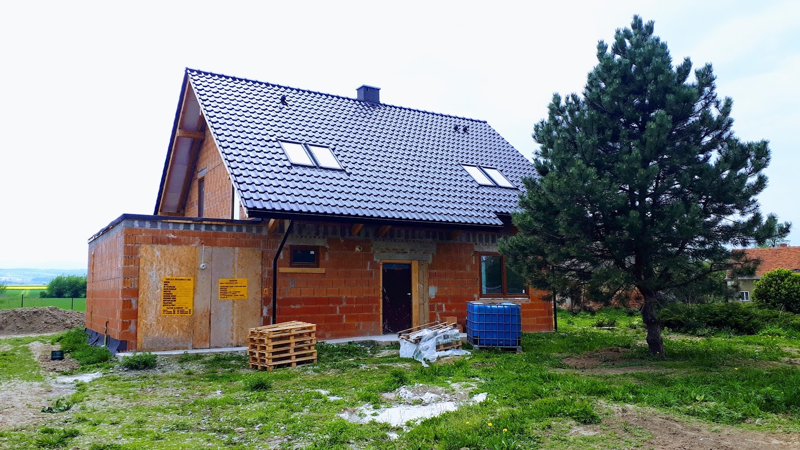 Surowy dom z czerwonej cegły z ciemnym dachem, okna dachowe, palety i niebieski pojemnik na trawniku. Budowa w toku, widok na wzgórza w tle.