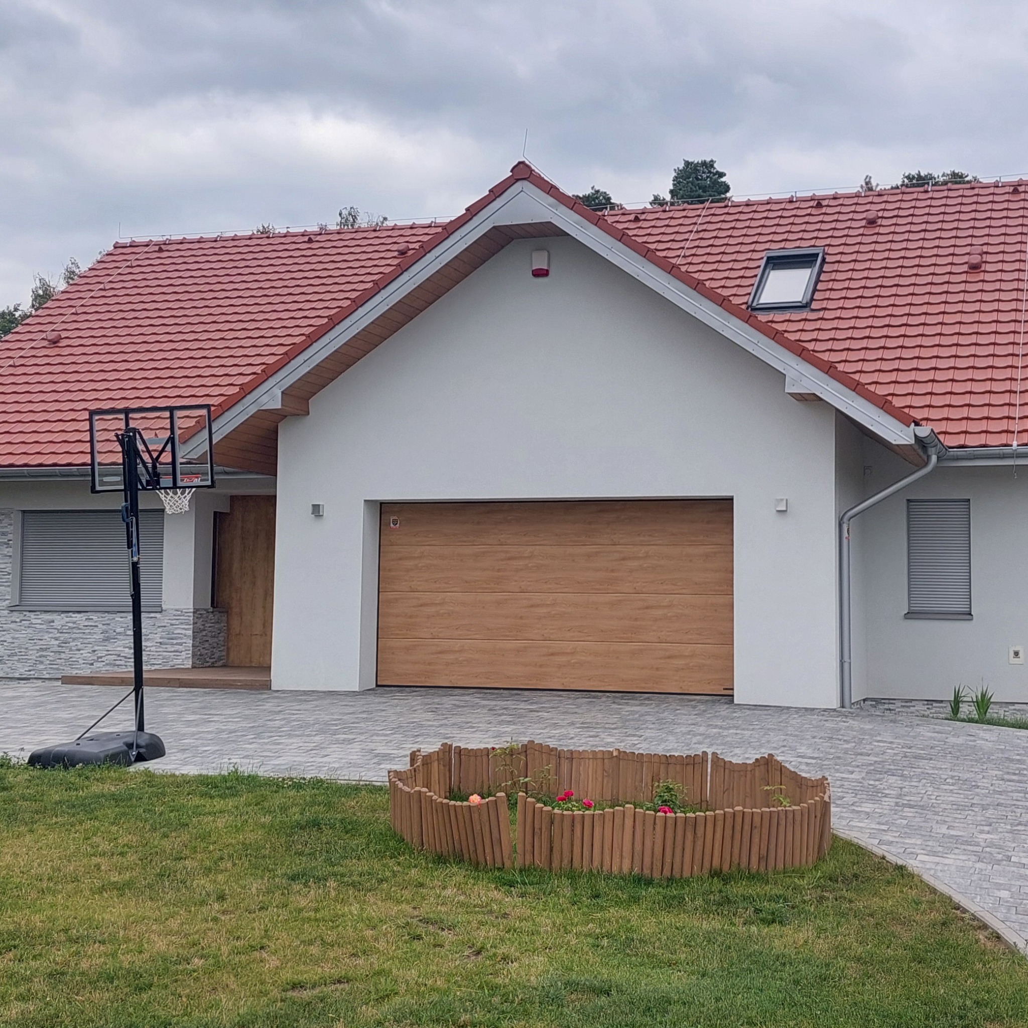 Nowoczesny dom jednorodzinny z garażem i czerwoną dachówką, widoczny z poziomu trawnika, z koszem do koszykówki po lewej stronie i drewnianym płotkiem rabaty kwiatowej.