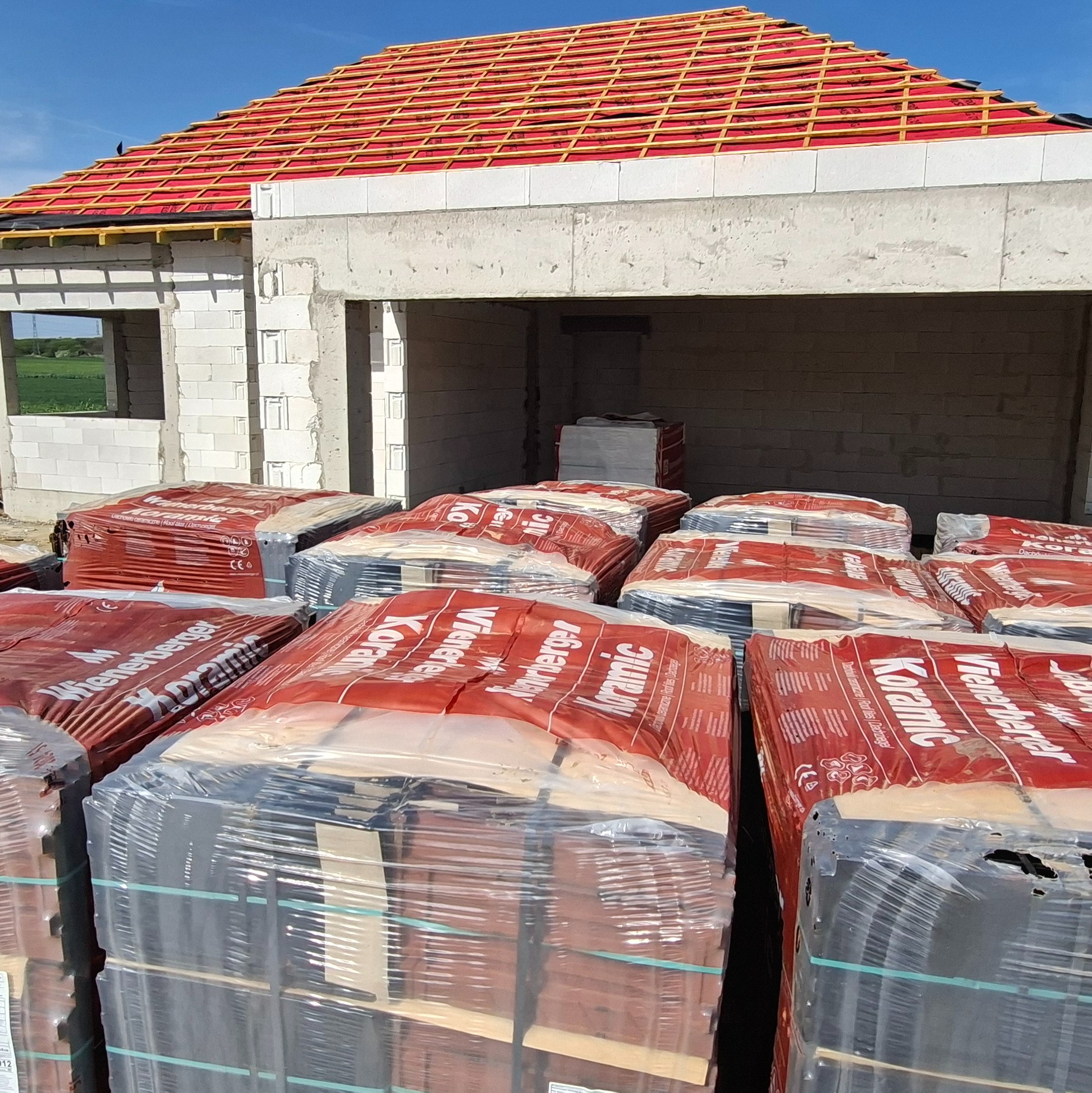 Budowa domu w trakcie realizacji: widoczne palety dachówek Koramic Wienerberger na tle surowej konstrukcji. Czerwona membrana dachowa pod łatami. Aerobetonowe ściany.