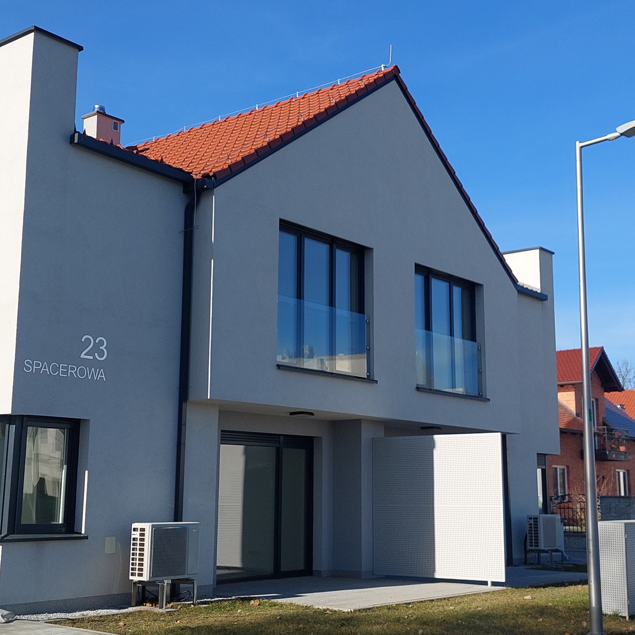 Nowoczesny dom jednorodzinny z szarą elewacją, pomarańczową dachówką i numerem '23 Spacerowa' na ścianie. Widoczne okna z balustradami i klimatyzatory na zewnątrz.