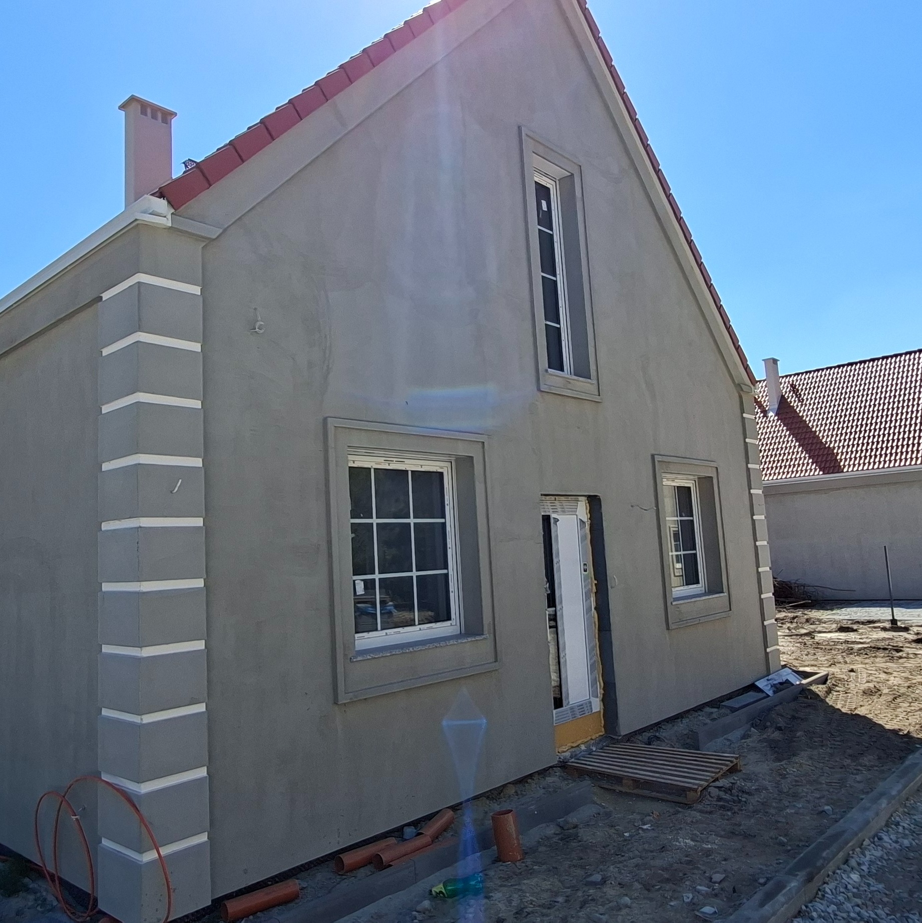 Szary dom jednorodzinny z czerwoną dachówką, wykończony dekoracyjnymi elementami elewacji, okna z białymi ramami. Widoczne rury i paleta przed budynkiem.