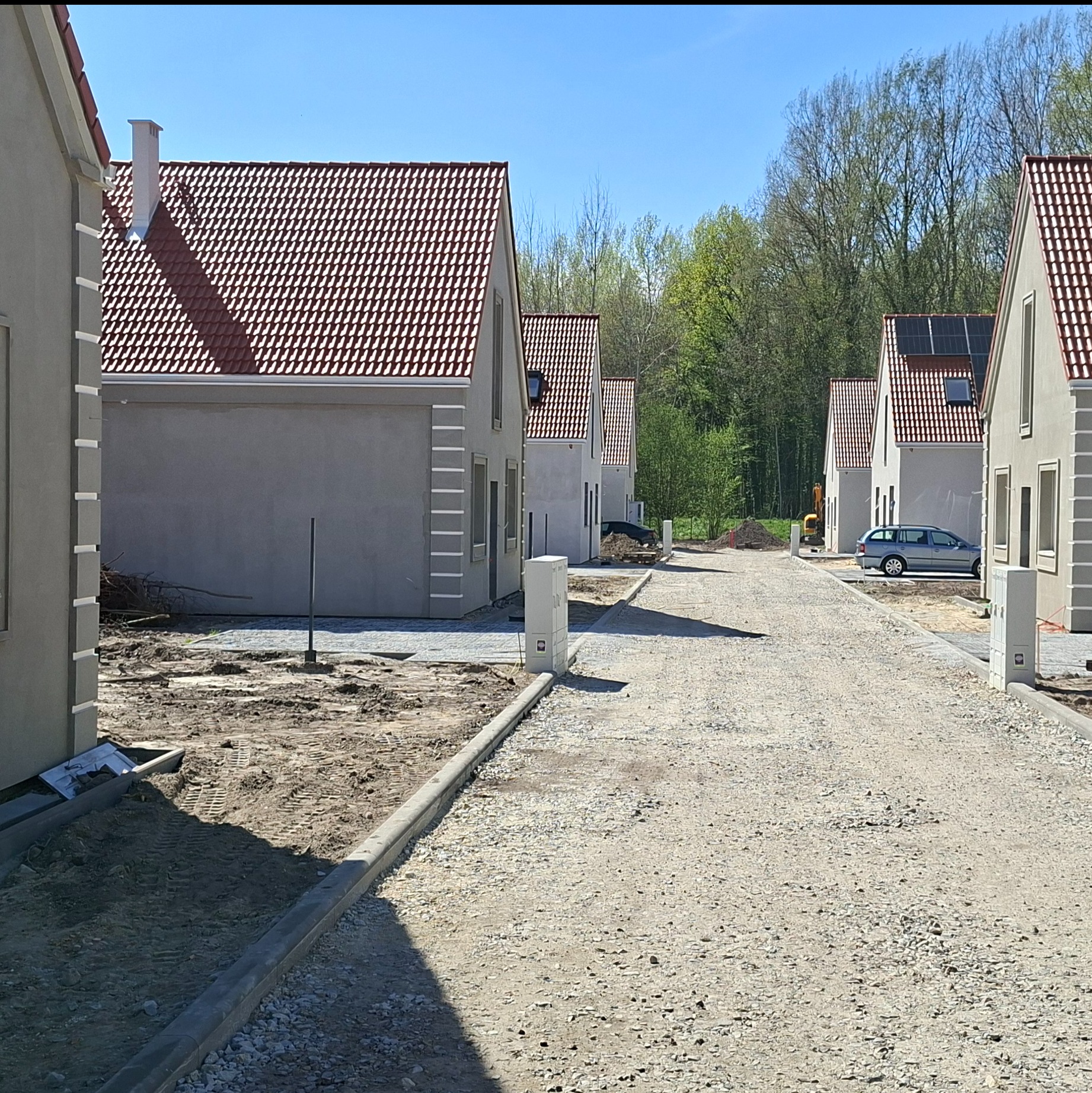 Nowoczesne domy jednorodzinne w zabudowie szeregowej z czerwoną dachówką i szarą elewacją, niektóre z panelami słonecznymi, na tle zieleni drzew.