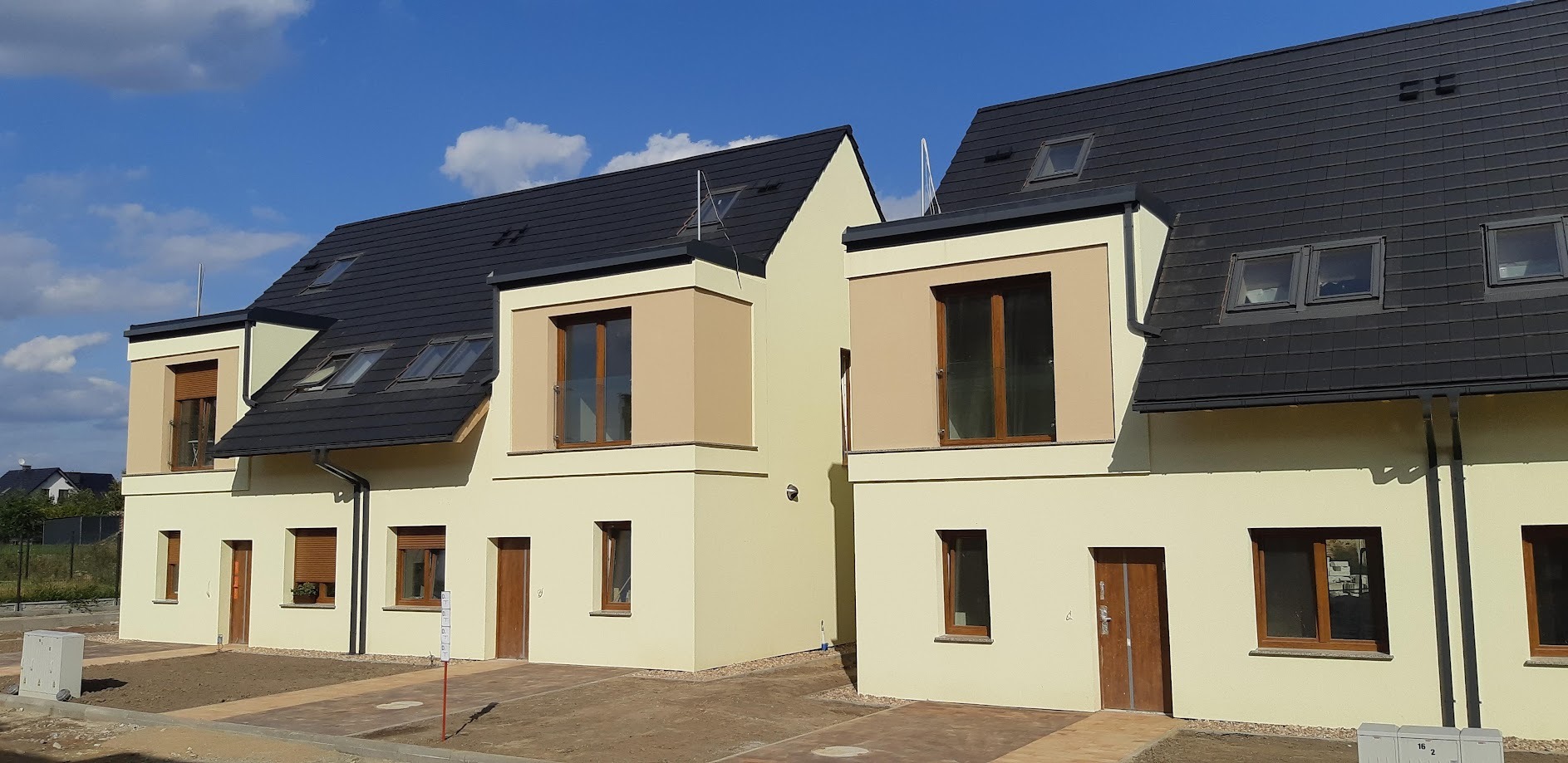 Nowoczesne domy bliźniacze z kremową elewacją i grafitowym dachem w słoneczny dzień. Widoczne okna dachowe i brązowe ramy okienne. Nieruchomość po budowie, teren uporządkowany.