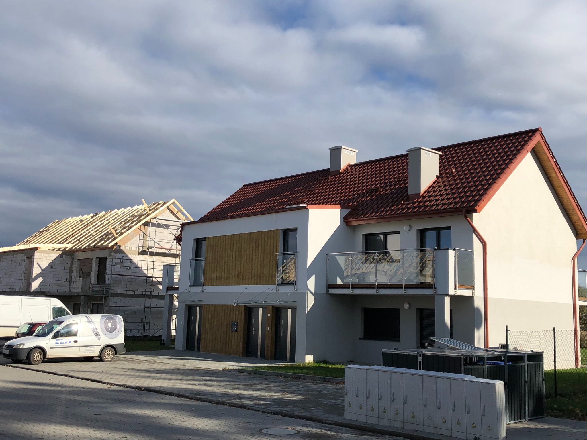 Nowoczesny dwupiętrowy dom z balkonem i czerwonym dachem, obok drugi dom w trakcie budowy z widoczną konstrukcją dachu, na pierwszym planie ulica i skrzynki elektryczne.