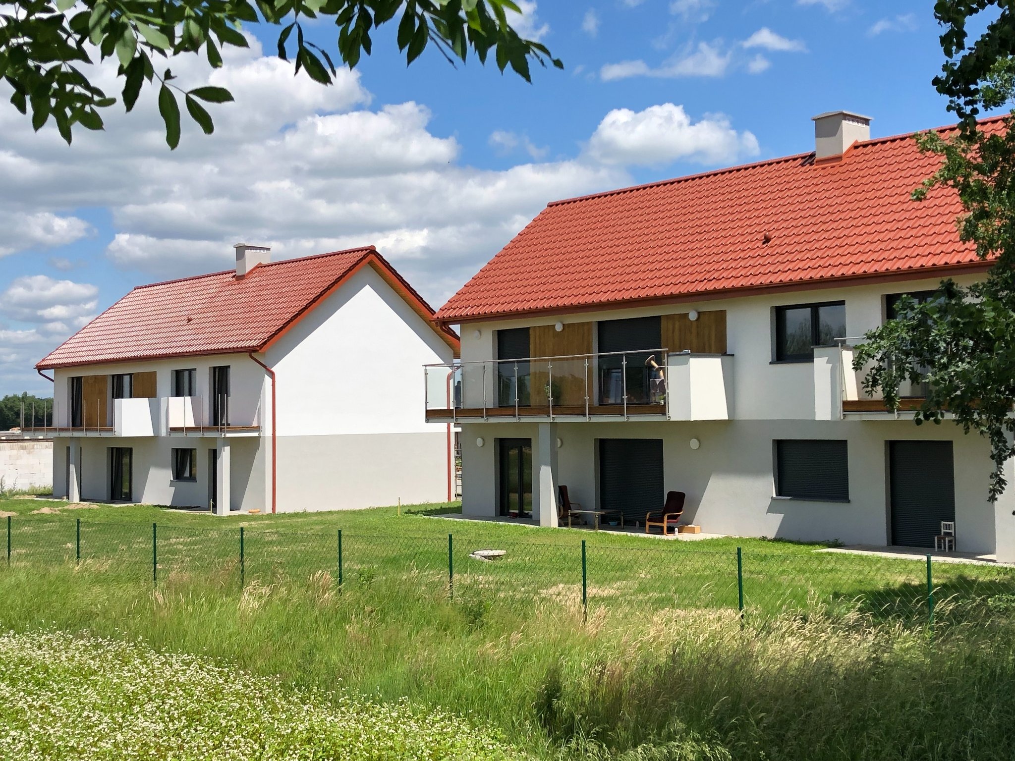 Dwa nowoczesne domy z czerwonymi dachami w słoneczny dzień. Widoczne balkony z szklanymi balustradami, białe ściany i zielone trawniki. Ujęcie zza ogrodzenia.