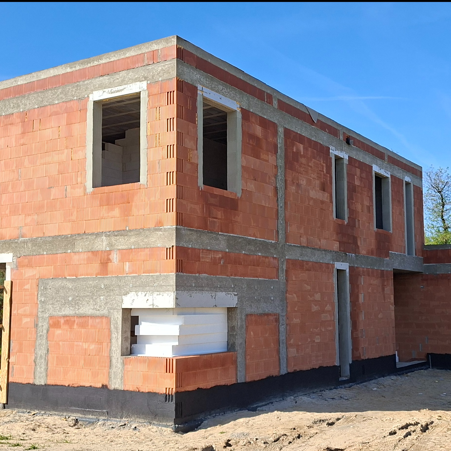 Dwukondygnacyjny dom w trakcie budowy z czerwonej cegły, z widocznym ociepleniem styropianem w dolnej części ściany i betonowymi nadprożami okiennymi, na tle błękitnego nieba.
