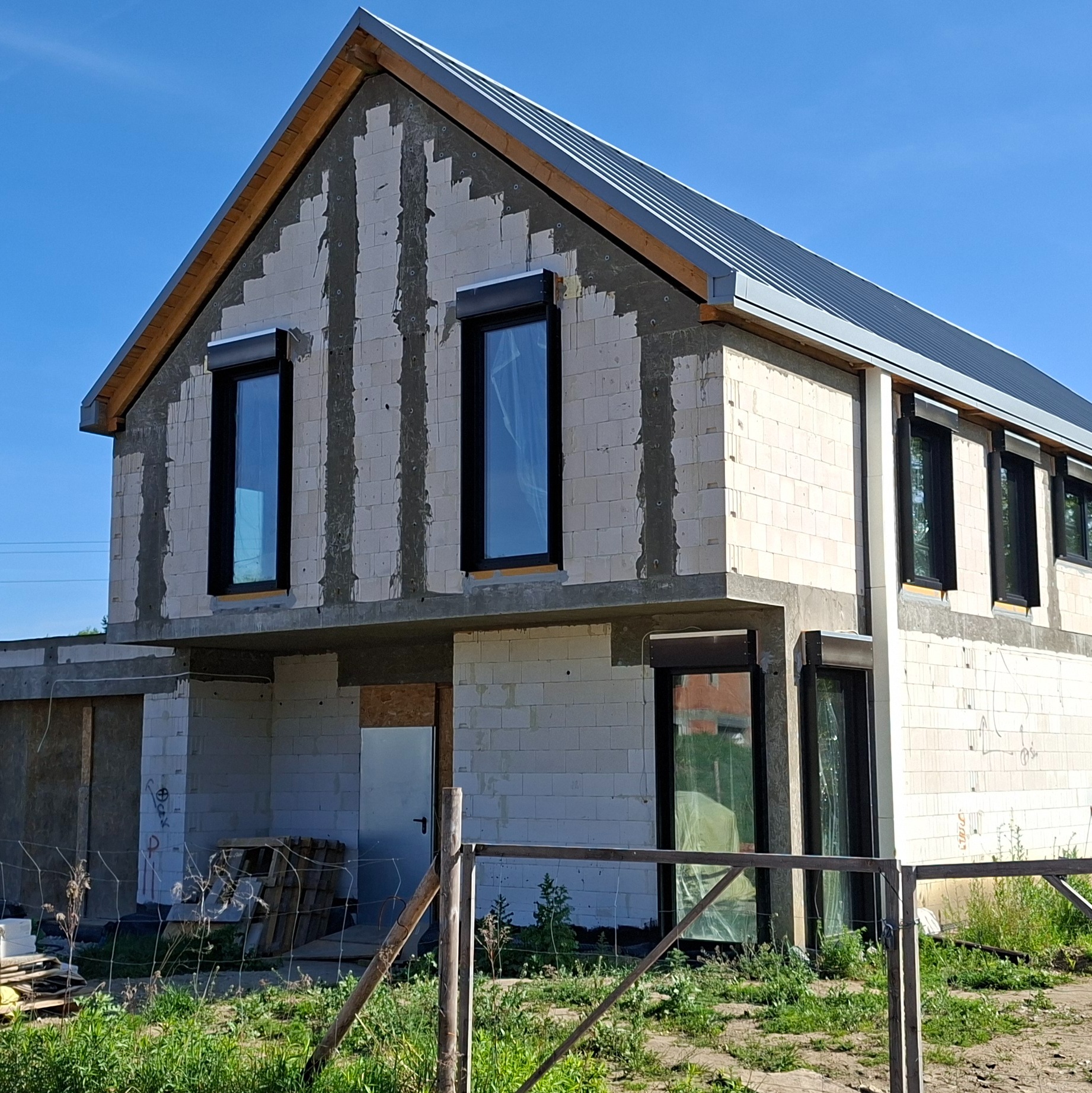 Dwukondygnacyjny dom w trakcie budowy, ściany z białych bloczków, czarne ramy okienne, szara blacha na dachu. Widoczne ślady po murowaniu.