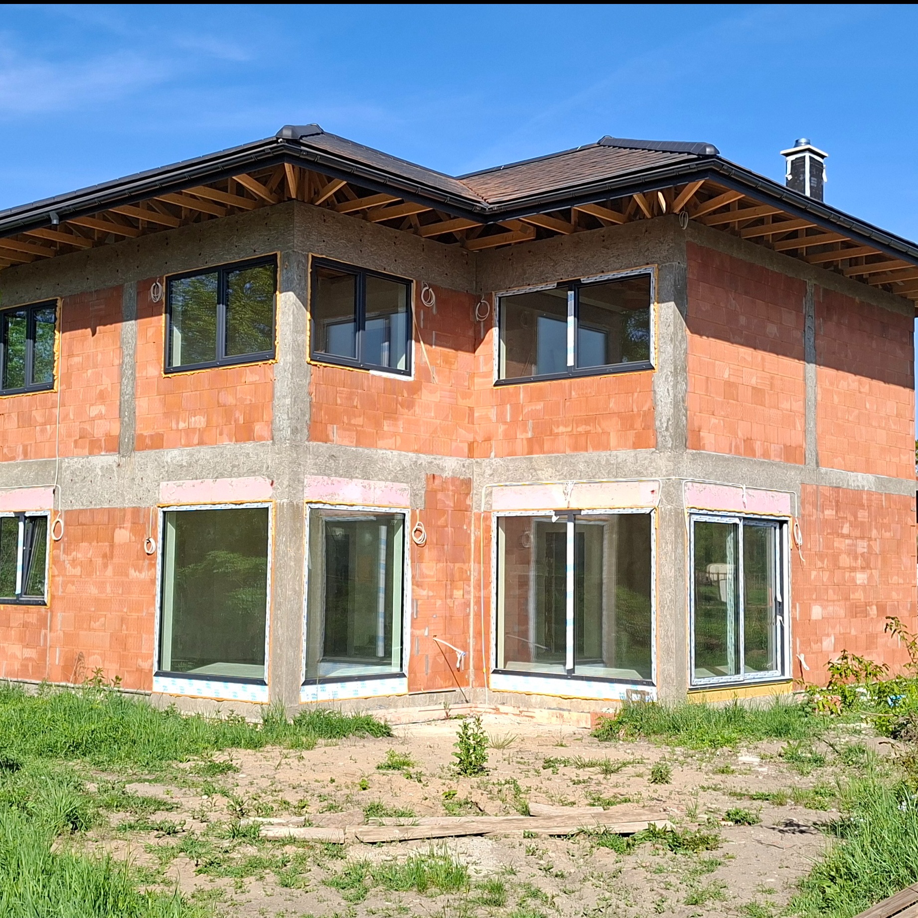 Dwukondygnacyjny dom w trakcie budowy, ściany z czerwonej cegły, czarne ramy okienne, widoczne elementy konstrukcji dachu, trawa i ziemia wokół budynku.