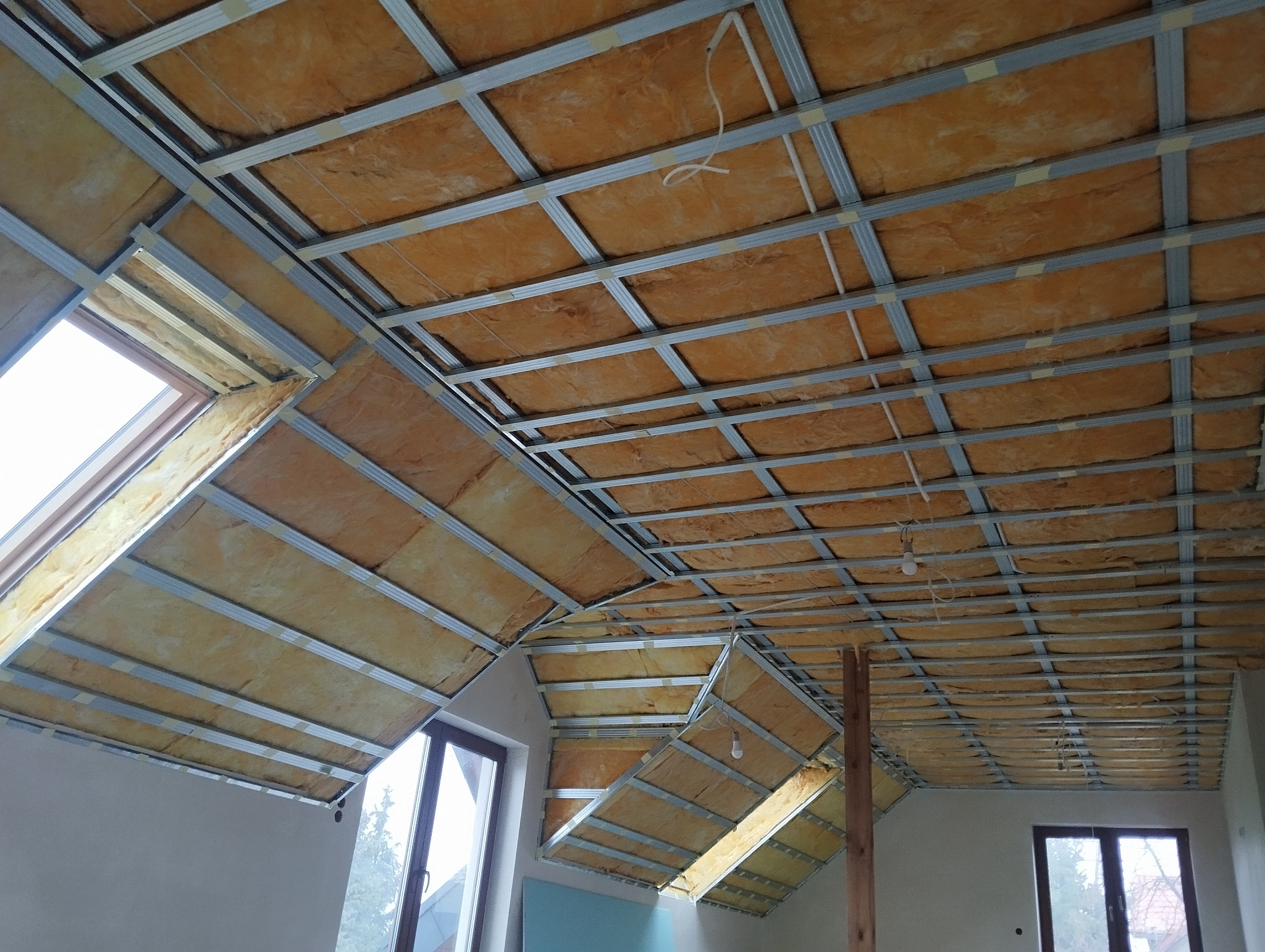 Sufit podwieszany z izolacją z wełny mineralnej w trakcie montażu. Widoczne metalowe profile i okna dachowe. Prace wykończeniowe na poddaszu.