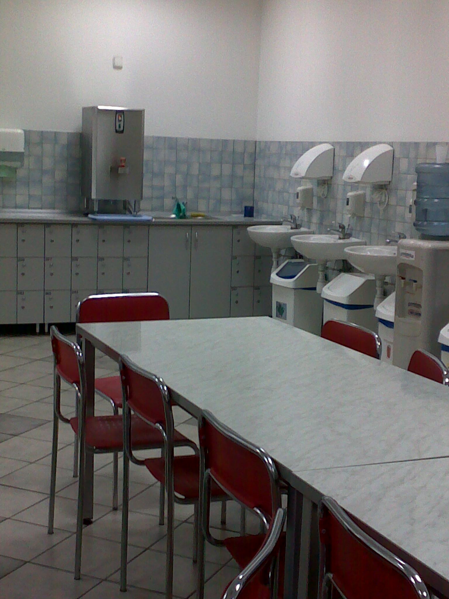 Stołówka zakładowa: długi stół z szarym blatem i czerwonymi krzesłami, w tle szafki pracownicze, umywalki z dozownikami mydła i automatyczne suszarki do rąk.