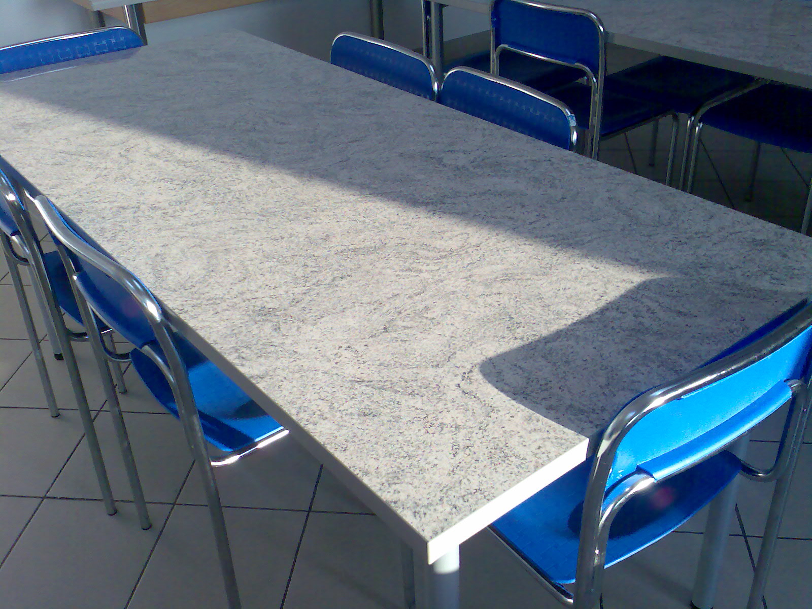 Długi stół z blatem imitującym granit, otoczony niebieskimi krzesłami z metalowymi nogami, oświetlony naturalnym światłem w jadalni.