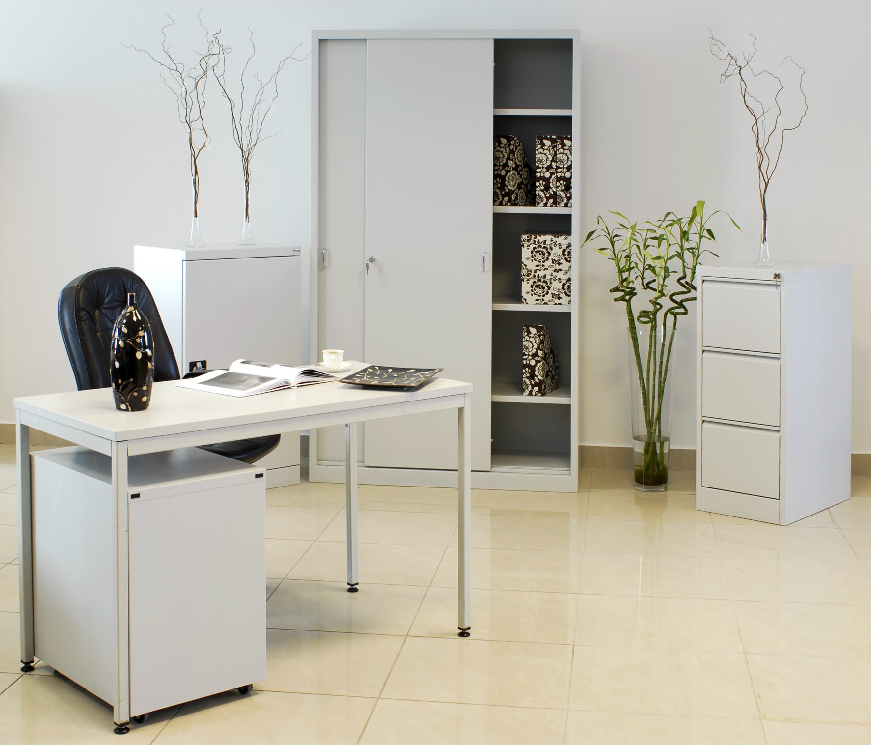 Białe meble biurowe: biurko z kontenerem, szafa przesuwna z półkami wypełnionymi pudełkami dekoracyjnymi, szafka kartotekowa, całość uzupełniona dekoracjami roślinnymi i wazonami z gałązkami.