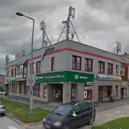 Panorama miejska z budynkiem banku z zielonym szyldem i antenami na dachu, pochmurne niebo w tle.