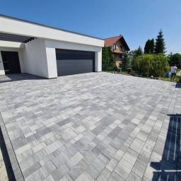 Geo-Bau Chmielarski - Szeroki podjazd z szarej kostki betonowej prowadzący do nowoczesnego garażu z ciemną bramą. W tle dom z czerwonym dachem i zieleń. Słoneczny dzień.