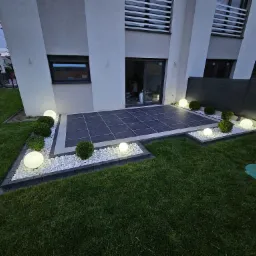 Nowoczesny taras z ciemnej kostki, otoczony białymi kamieniami i kulistymi lampami, przylegający do budynku z grafitowymi oknami i zielonym trawnikiem.