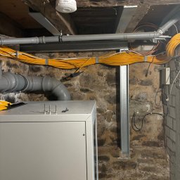 Geo-Bau Chmielarski - Instalacja elektryczna w piwnicy z rurami PCV i metalowymi wspornikami. Widoczne przewody w żółtej izolacji oraz elementy centrali. Kamienna ściana w tle.