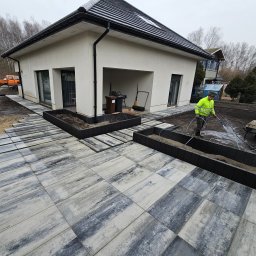 Geo-Bau Chmielarski - Układanie kostki brukowej na posesji przy domu. Pracownik w żółtej kurtce spryskuje powierzchnię wodą. Widoczne rabaty i elementy małej architektury.