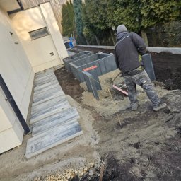 Geo-Bau Chmielarski - Układanie betonowych obrzeży rabat w ogrodzie. W tle widoczny pracownik z łopatą. Częściowo ułożony chodnik z płyt betonowych przy budynku.