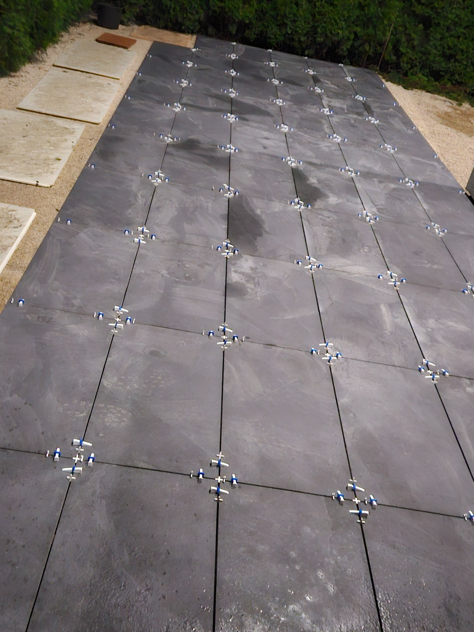 Układanie ciemnoszarej kostki brukowej na zewnątrz, z użyciem krzyżyków dystansowych, widok z góry, zieleń w tle. Kostka częściowo ułożona na piasku.