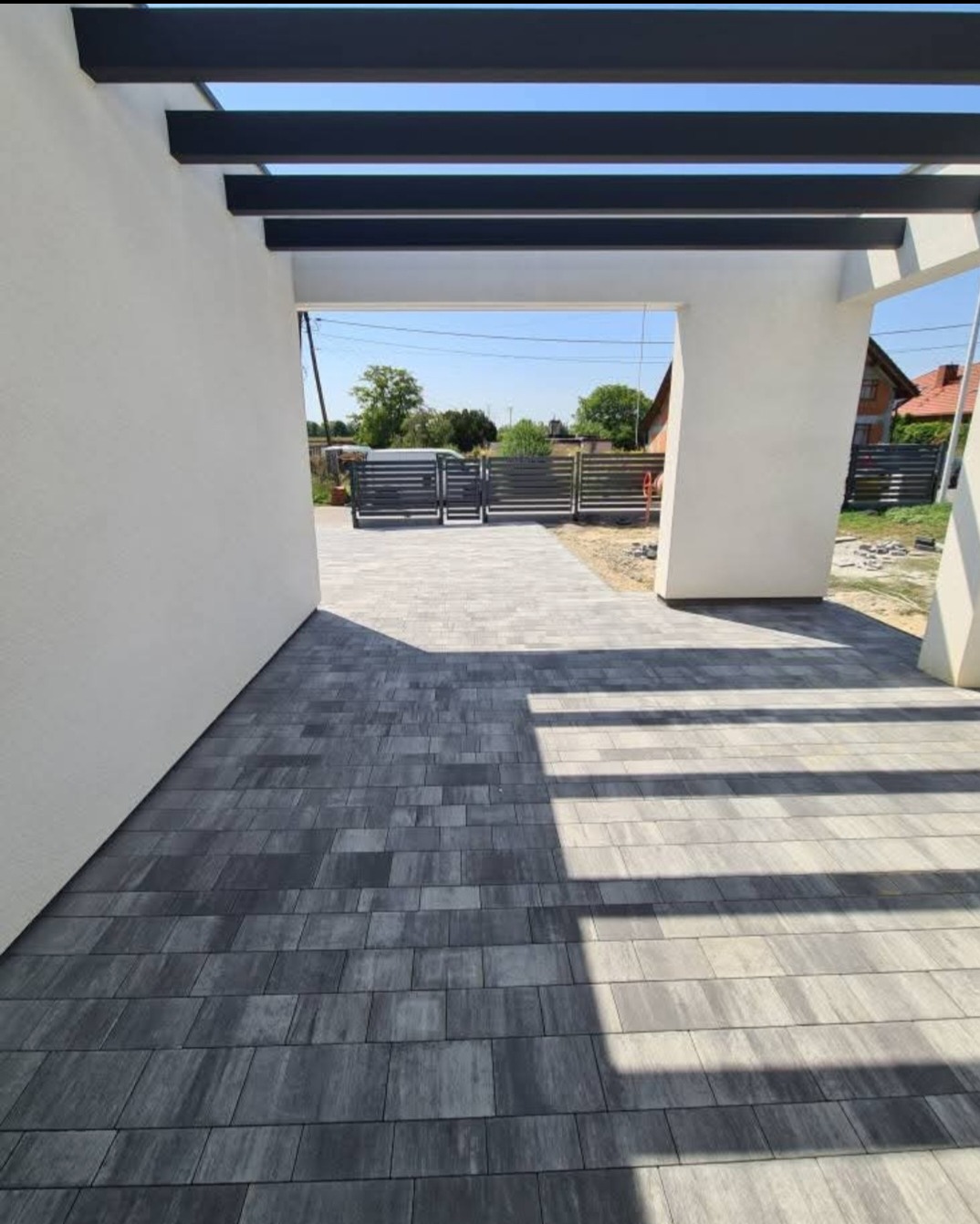 Ułożona kostka betonowa w kolorze szarości prowadząca do bramy wjazdowej. Widoczne elementy konstrukcyjne z ciemnego drewna. Słoneczny dzień.