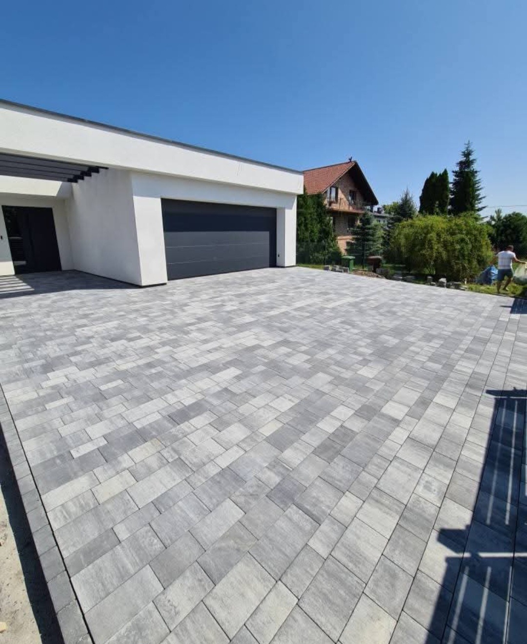 Szeroki podjazd z szarej kostki betonowej prowadzący do nowoczesnego garażu z ciemną bramą. W tle dom z czerwonym dachem i zieleń. Słoneczny dzień.