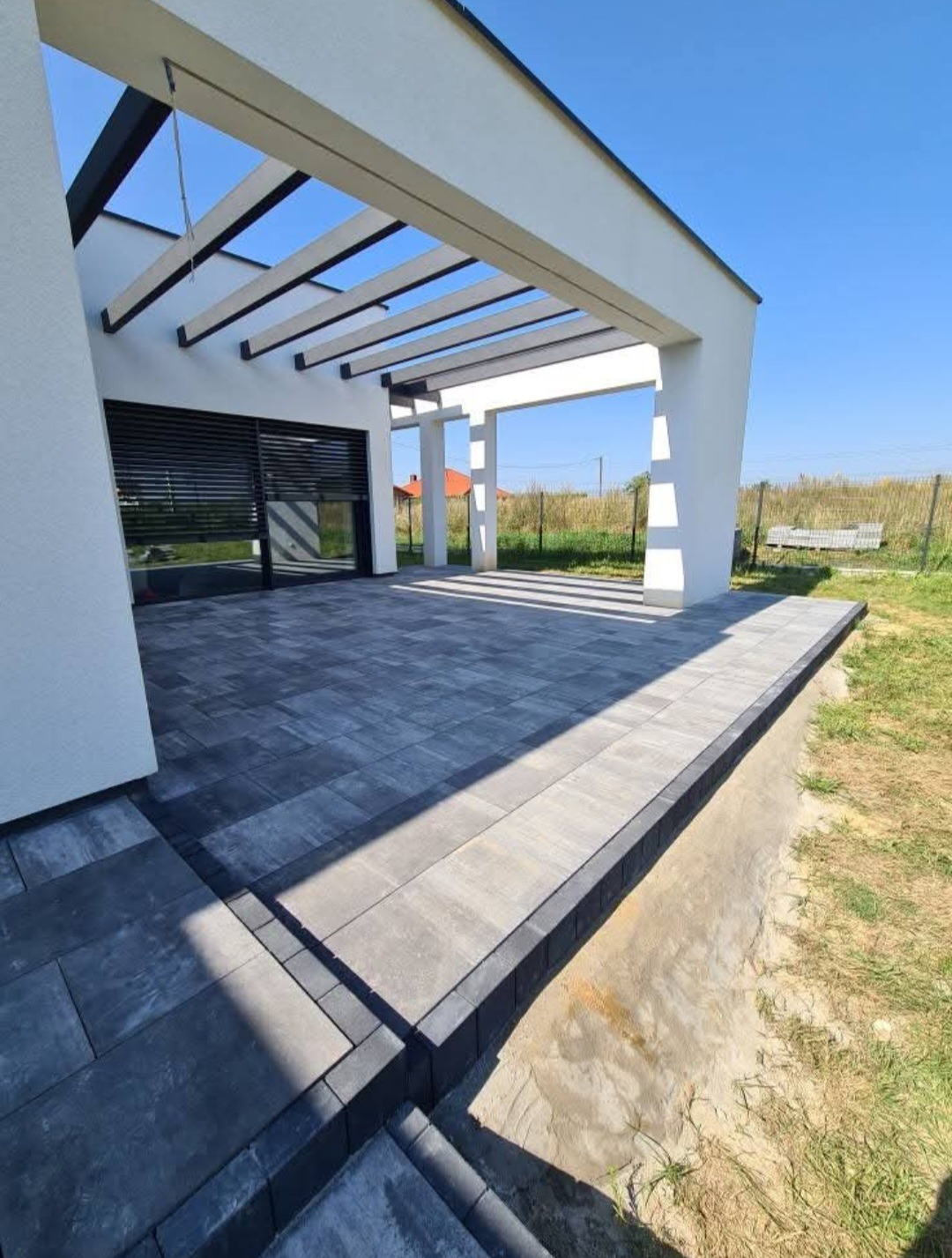 Nowoczesny taras z szarej kostki betonowej, częściowo zadaszony białą konstrukcją. Widoczne subtelne cieniowanie i staranne ułożenie nawierzchni.