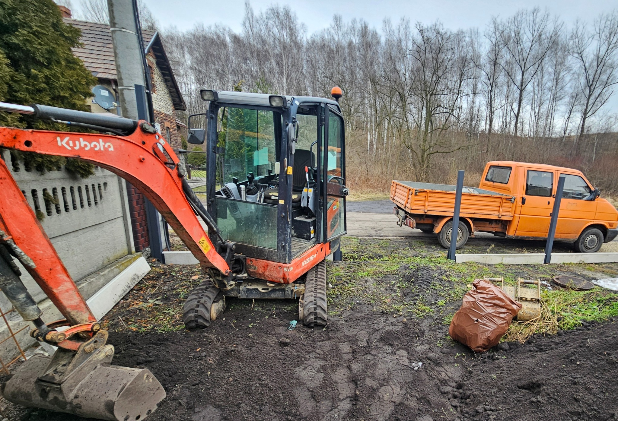 Pomarańczowa koparka Kubota stoi na błotnistym terenie obok ogrodzenia i pomarańczowego samochodu dostawczego. W tle budynek i drzewa.