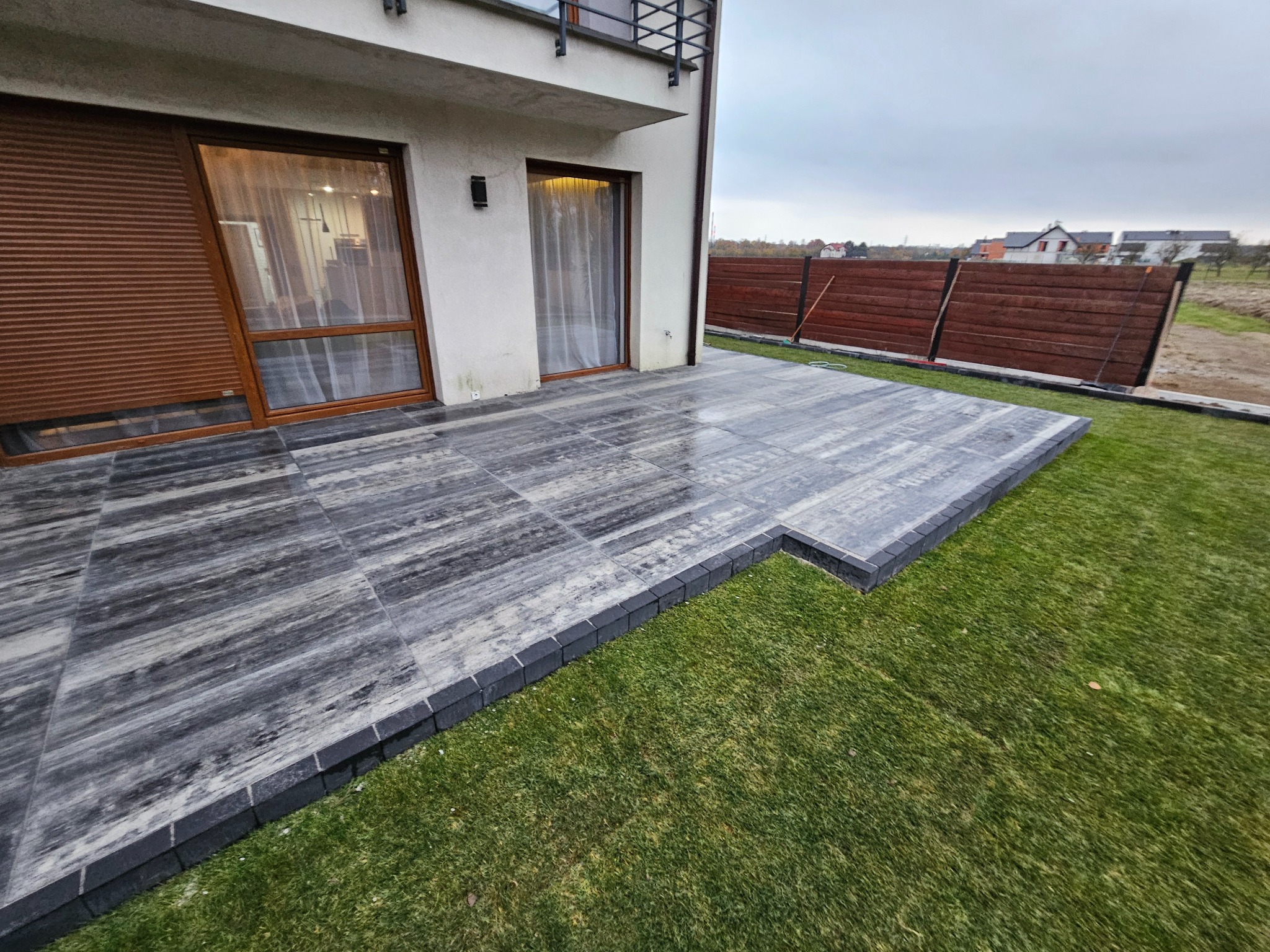 Nowoczesny taras z szarych płyt imitujących drewno, obramowany ciemną kostką, przylegający do domu z brązowymi roletami i drewnianym ogrodzeniem w tle.