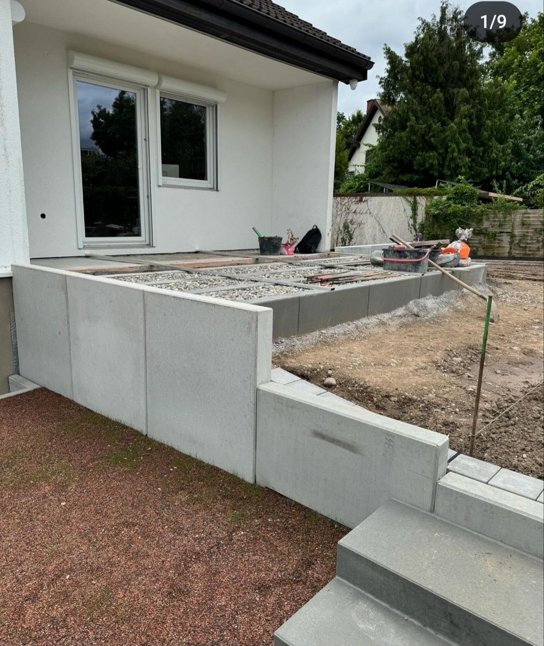 Budowa tarasu z betonowych elementów: podmurówka, schody i podsypka z kruszywa. Widoczne narzędzia i materiały budowlane. Prace w toku.