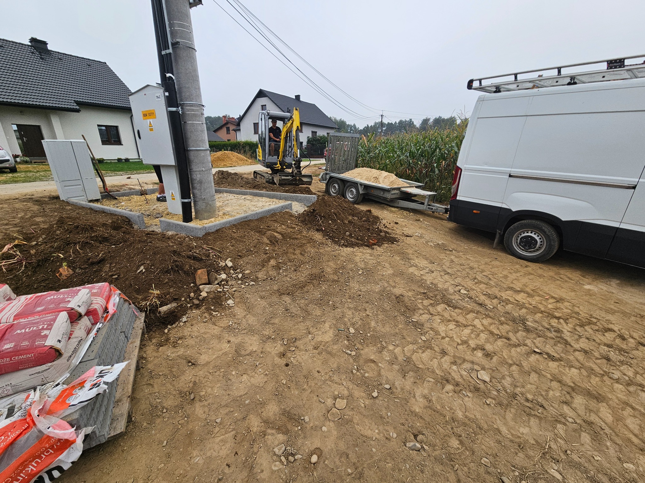 Prace ziemne przy układaniu obrzeży z kostki w Chełm Śląski. Mini koparka i przyczepka z piaskiem w tle. Paleta z cementem i kostką na pierwszym planie.