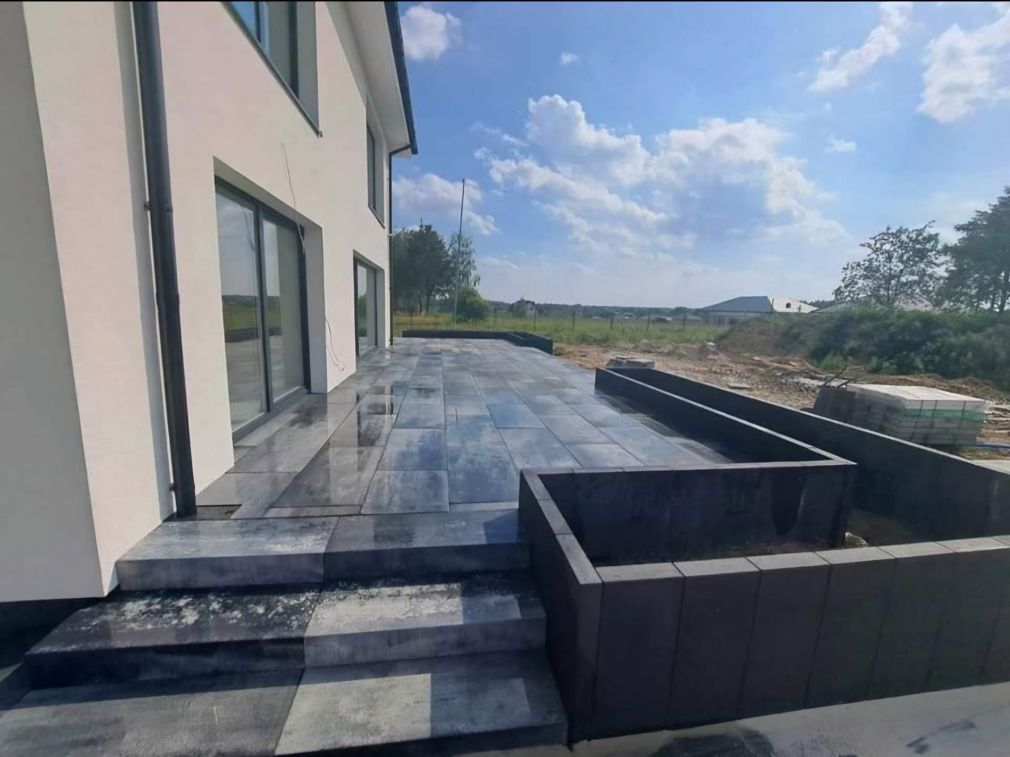 Nowoczesny taras z szarej kostki betonowej przy domu z dużymi oknami, w tle błękitne niebo z chmurami. Widoczne schody i obrzeża rabat z kostki.