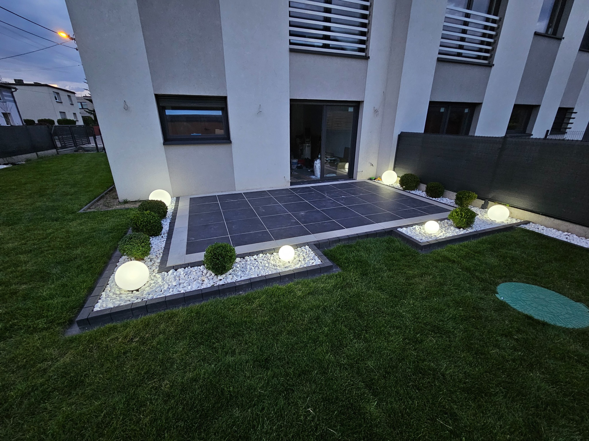 Nowoczesny taras z ciemnej kostki, otoczony białymi kamieniami i kulistymi lampami, przylegający do budynku z grafitowymi oknami i zielonym trawnikiem.