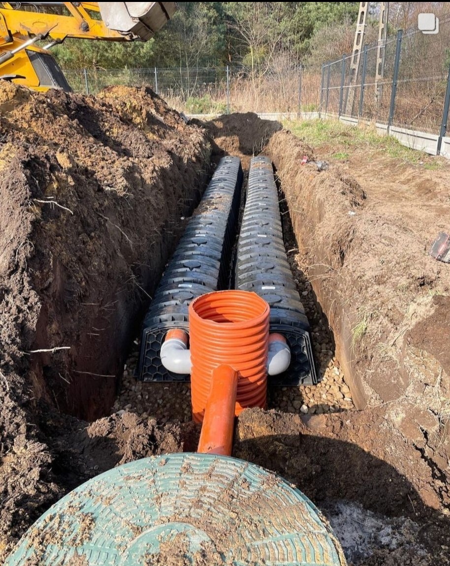 Drenaż rozsączający w wykopie, z widocznymi rurami drenarskimi i studzienką kontrolną. Częściowo zasypane żwirem. W tle widoczne drzewa i ogrodzenie.