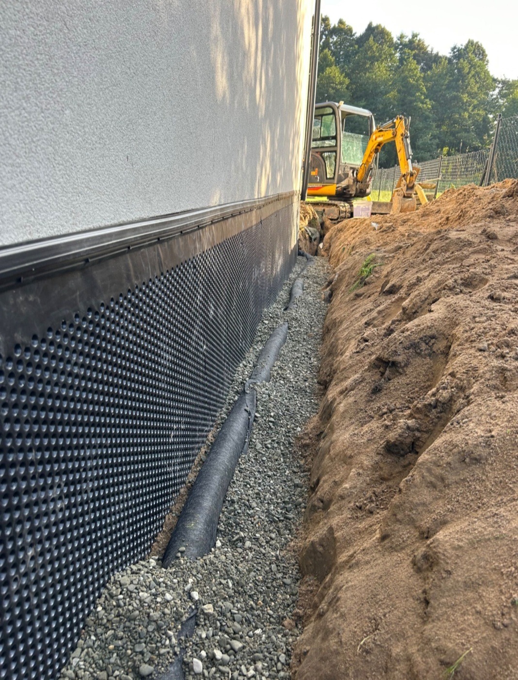 Ocieplenie fundamentów budynku z folią kubełkową, drenażem i warstwą żwiru. W tle koparka. Budowa fundamentów w trakcie realizacji.