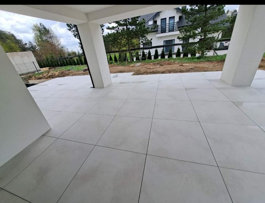 Jasnoszara kostka na tarasie domu jednorodzinnego z widokiem na ogród i budynek w tle. Minimalistyczny design i nowoczesna estetyka.