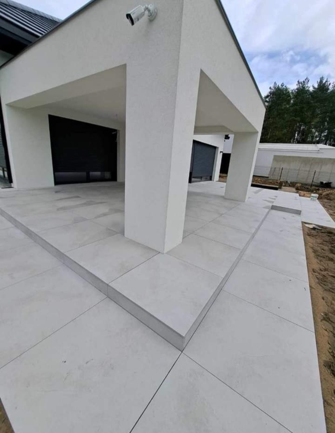 Nowoczesny dom z białymi kolumnami i szarą kostką brukową. Widoczny system rolet zewnętrznych i kamera monitoringu. Minimalistyczny design z elementami architektury kubistycznej.