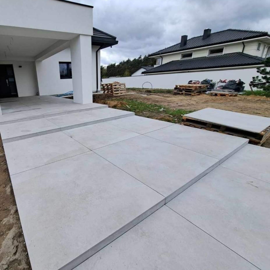 Nowoczesny podjazd z jasnych, kwadratowych płyt betonowych prowadzący do domu. Widoczne palety z materiałami budowlanymi w tle.